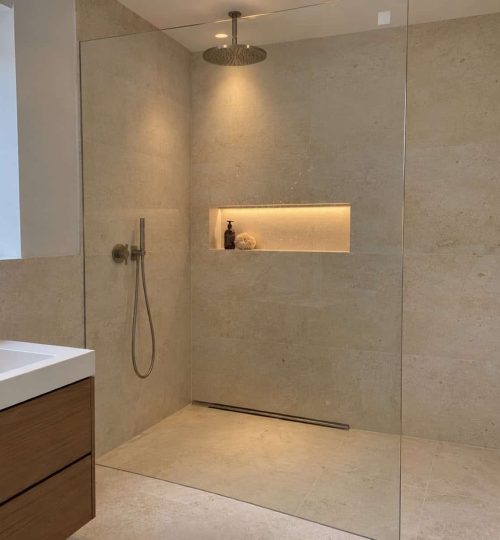Bodengleiche Dusche in cremefarbenem Kalkstein mit Regenbrause an der Decke, einer beleuchteten Wandnische und einer Ablaufrinne direkt an d